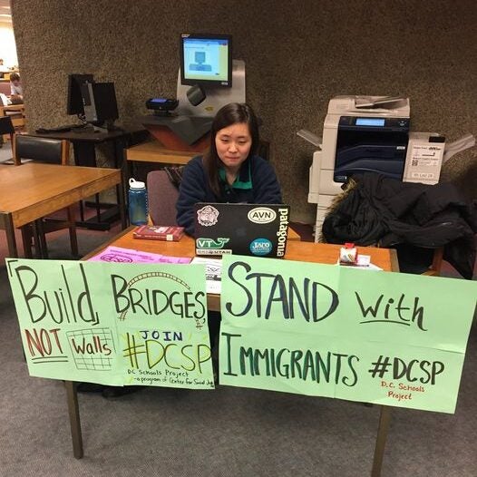 Student sits at a desk with informative signs about SIRA stating "Build Bridges Not Walls" and "Stand with Immigrants."