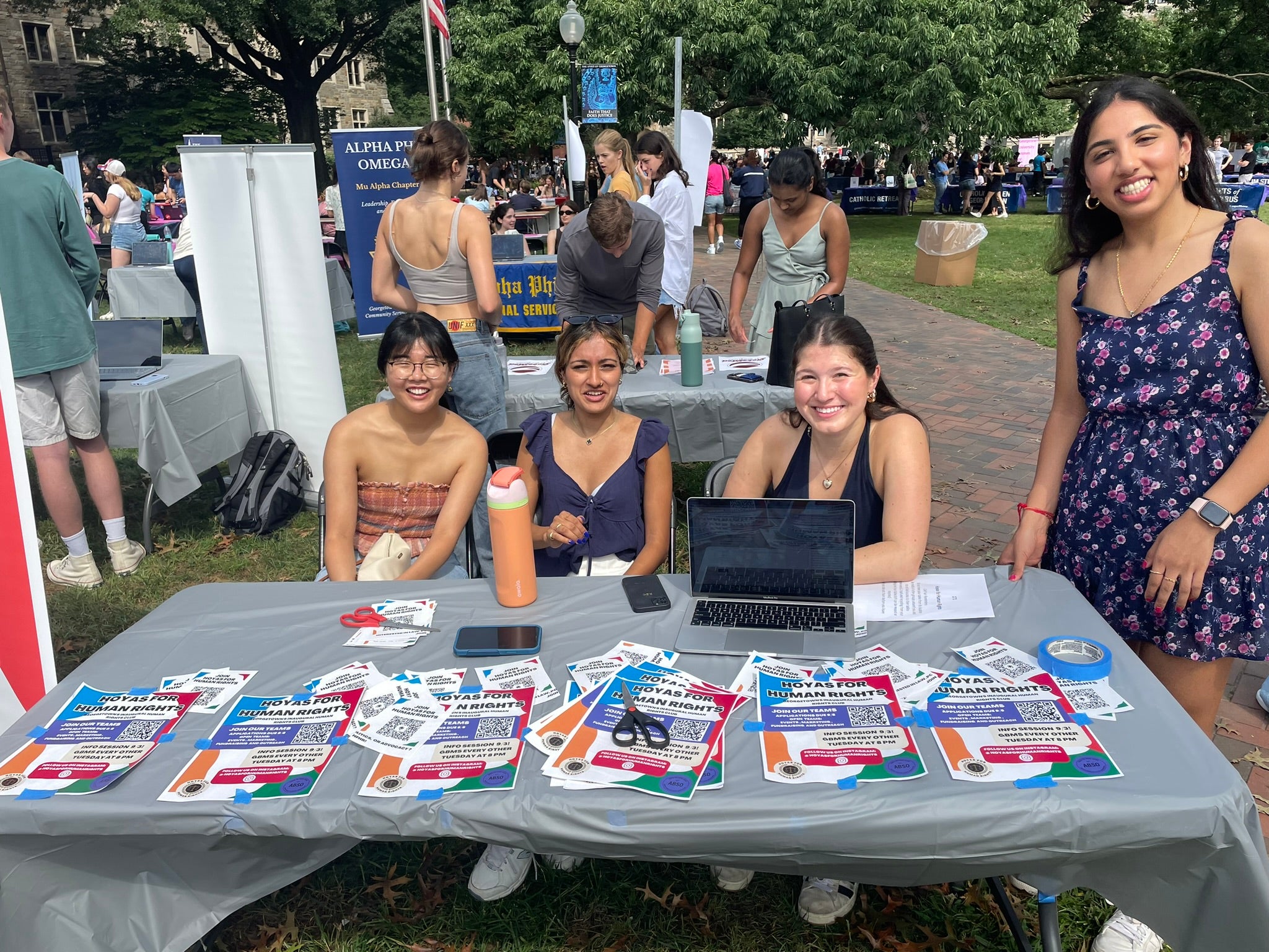 Three women sitting and one woman standing outside at a table full of flyers on a law full of tables and people