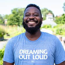 man smiling big at the camera wearing a tshirt that says Dreaming Out Loud