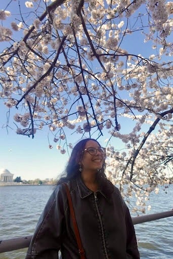 woman smiling wearing glasses under a branch of pink and white cherry blossoms