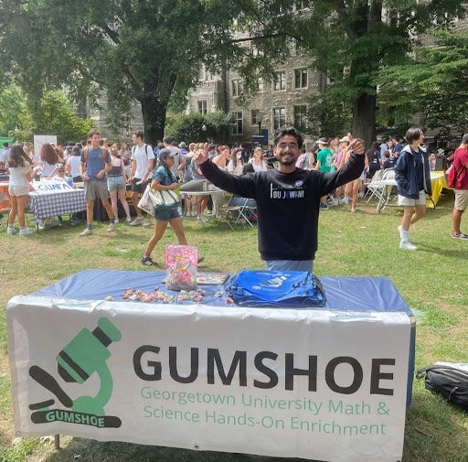 A man stands behind a table displaying a sign that reads "Gumshoe," indicating a possible event or exhibit. He has his arms raised and he looks excited.